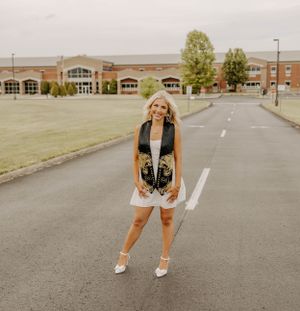 Graduate poses on campus road wearing black graduation gown and white dress.