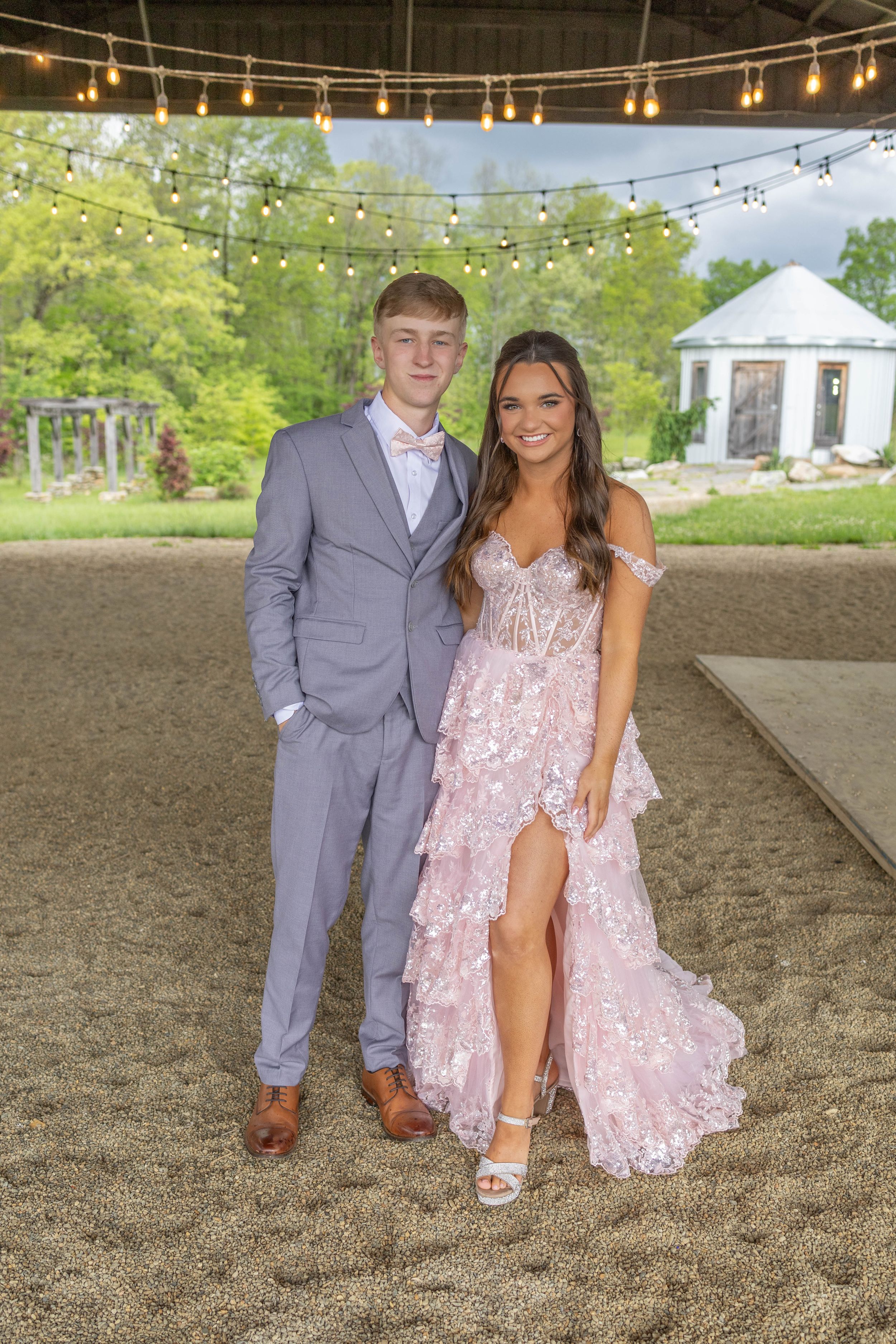 A formal couple poses under string lights in elegant prom attire.