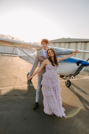 Couple in formal wear poses playfully in front of small aircraft at sunset.