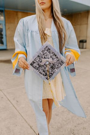 Graduate holds decorated graduation cap while wearing light blue graduation gown.