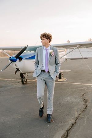 Young man in light gray suit and purple tie poses in front of small airplane.