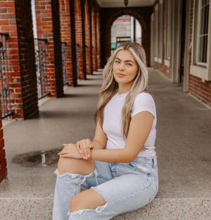 Graduate in casual attire sits in covered walkway with brick columns.