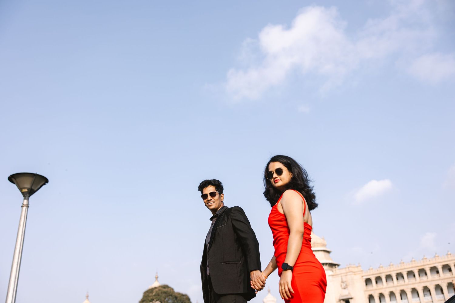 Stylish couple poses against blue sky with a streetlamp and building in background.