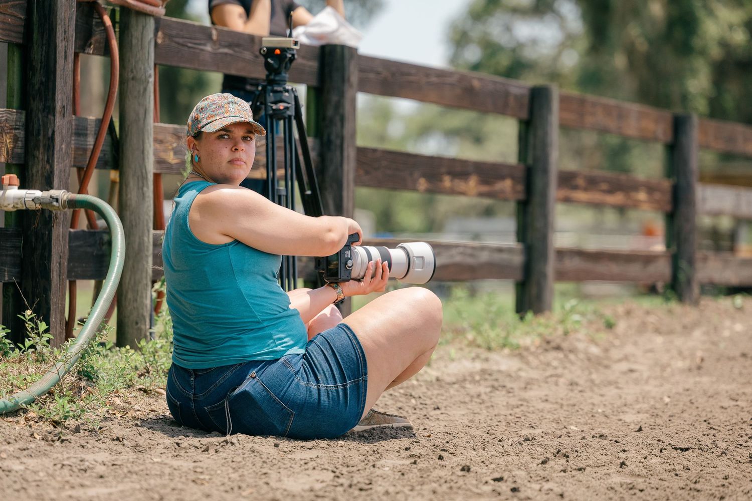 A photographer in a teal tank top and denim shorts sits on the ground with camera equipment near a wooden fence.