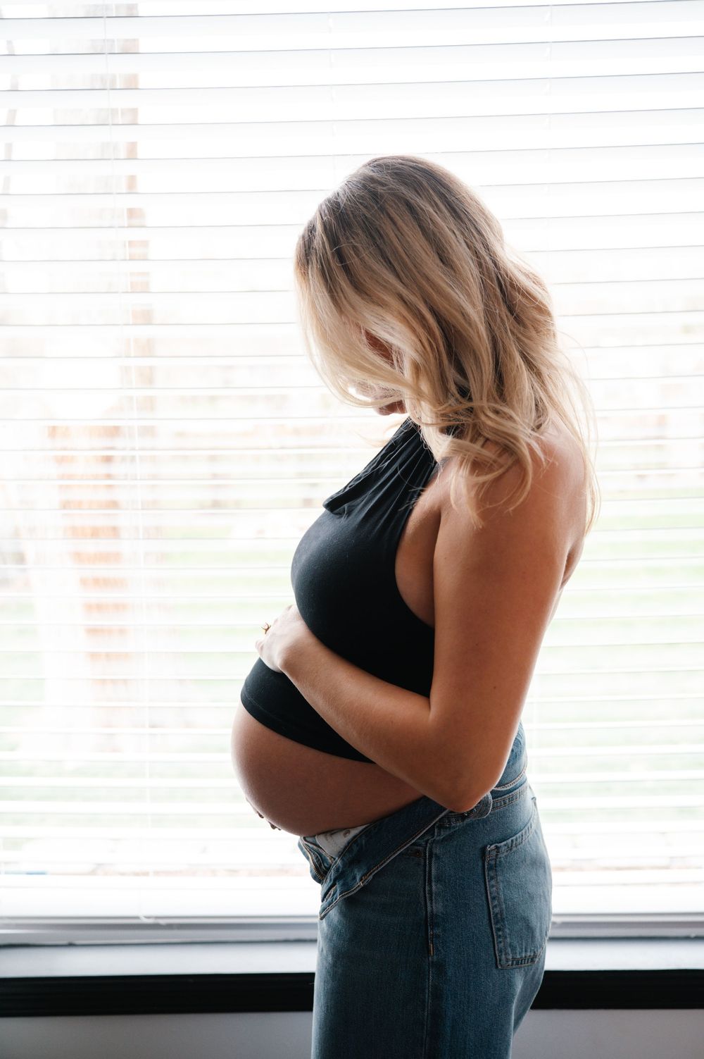 Pregnant woman in black crop top and jeans holding baby bump while standing by a sunlit window.