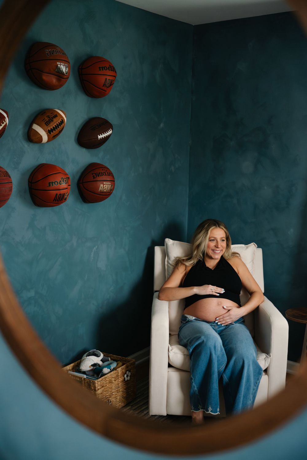 Smiling pregnant woman sitting in white nursery chair surrounded by sports balls on teal wall.