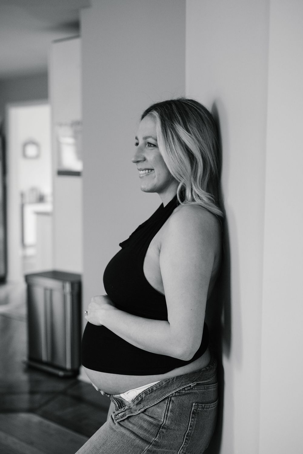 Black and white photo of pregnant woman in black top leaning against wall smiling sideways.