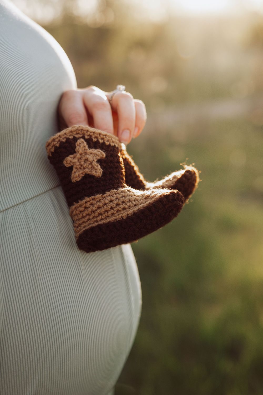 Pregnant woman holding crocheted brown cowboy boots against her baby bump in sunlight.