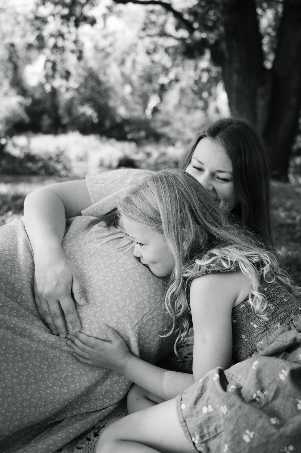 Black and white photo of young girl hugging pregnant mothers belly outdoors under trees.