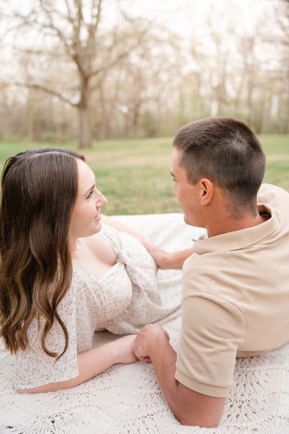 Couple lying on white blanket in park, gazing at each other during maternity session.