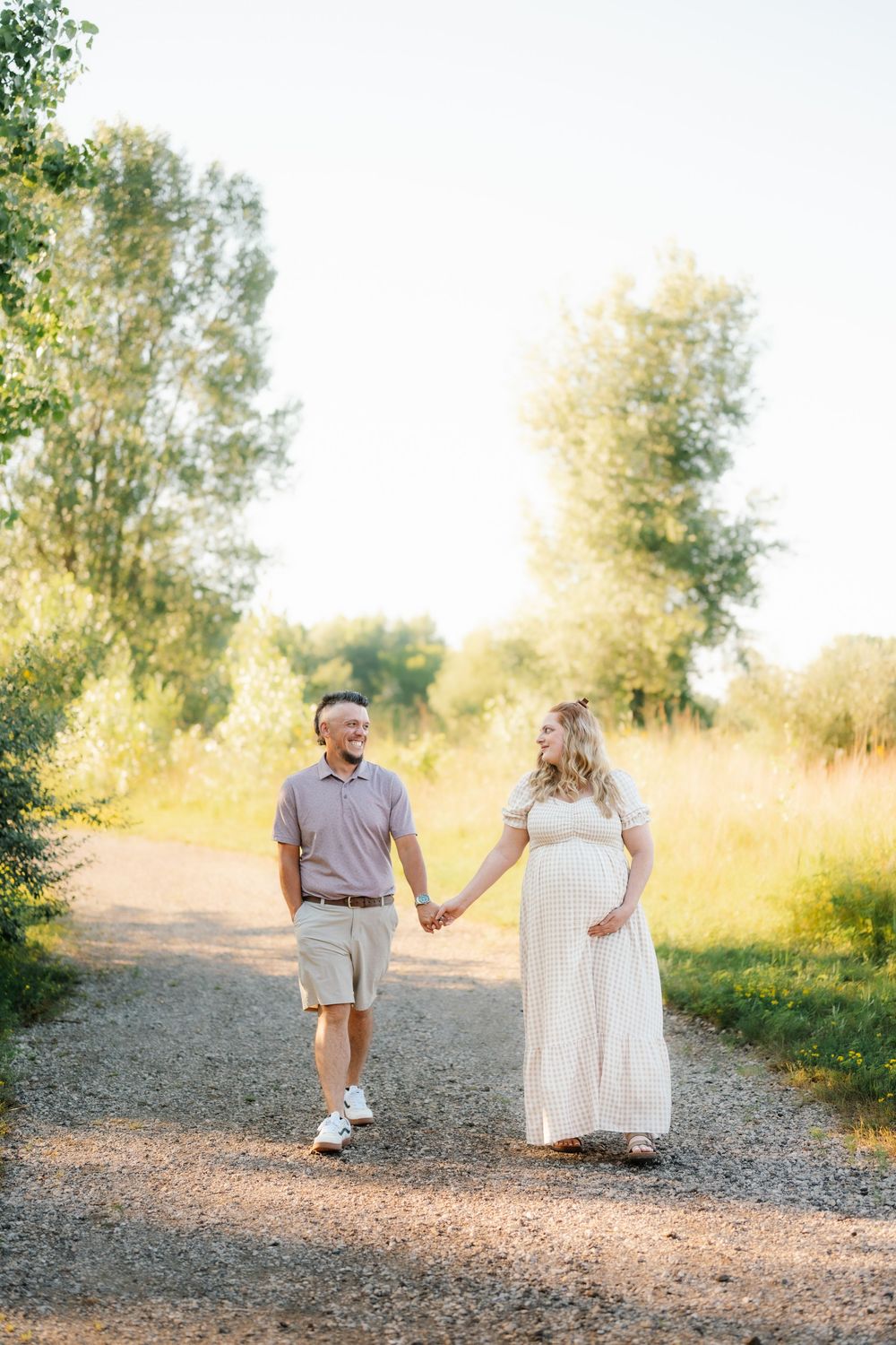 Pregnant couple holding hands walking on a sunny gravel path surrounded by lush green trees during golden hour.