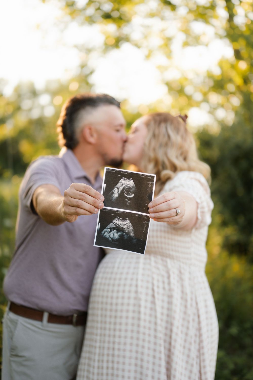 Couple kissing outdoors while holding ultrasound photos, announcing pregnancy in a sunny green nature setting.