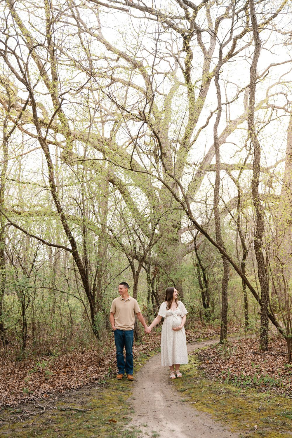 Pregnant couple holding hands walking on forest path surrounded by moss-covered trees.