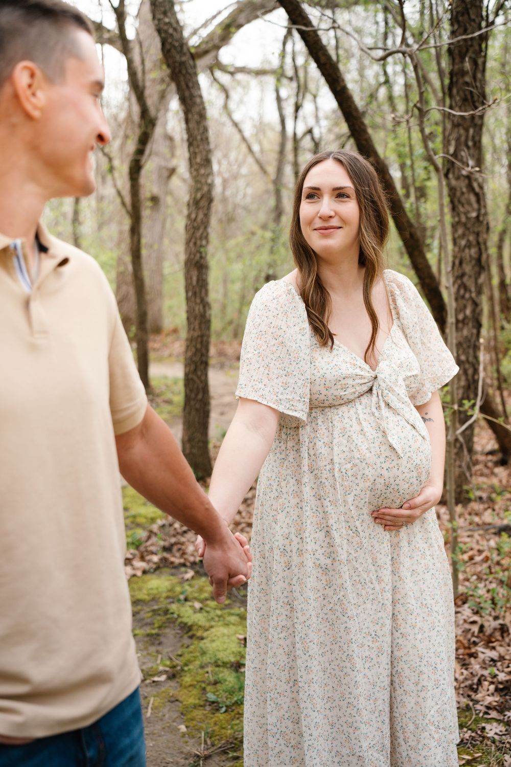 Pregnant woman in floral dress holding baby bump while holding hands with partner in woods.