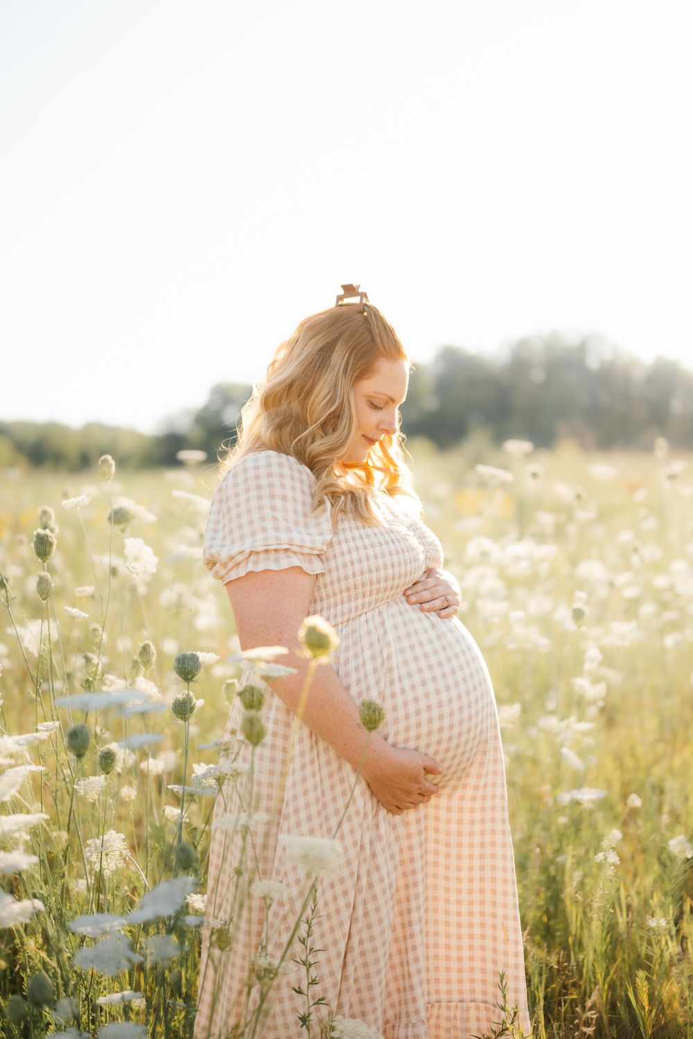 Pregnant woman in gingham dress cradling belly in sunlit wildflower field during golden hour.