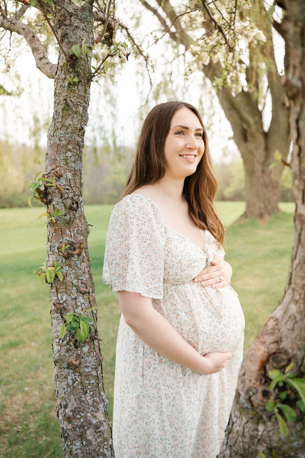 Smiling pregnant woman in floral dress cradling baby bump outdoors near blooming trees in spring.