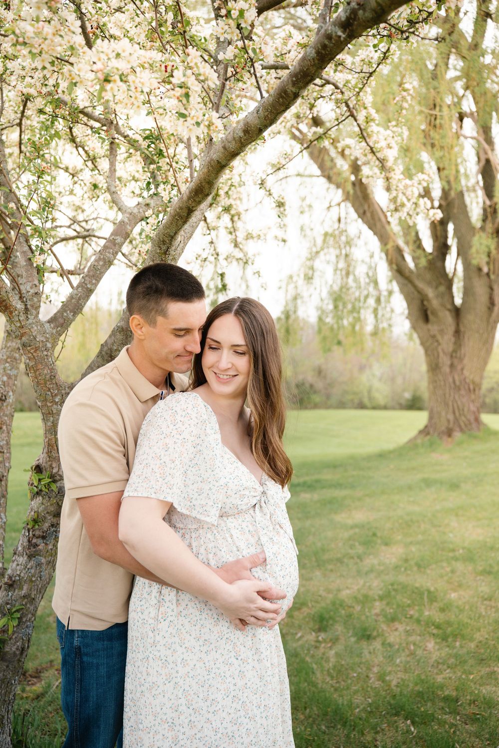 Happy couple embracing outdoors under flowering trees, woman in floral maternity dress holding baby bump.