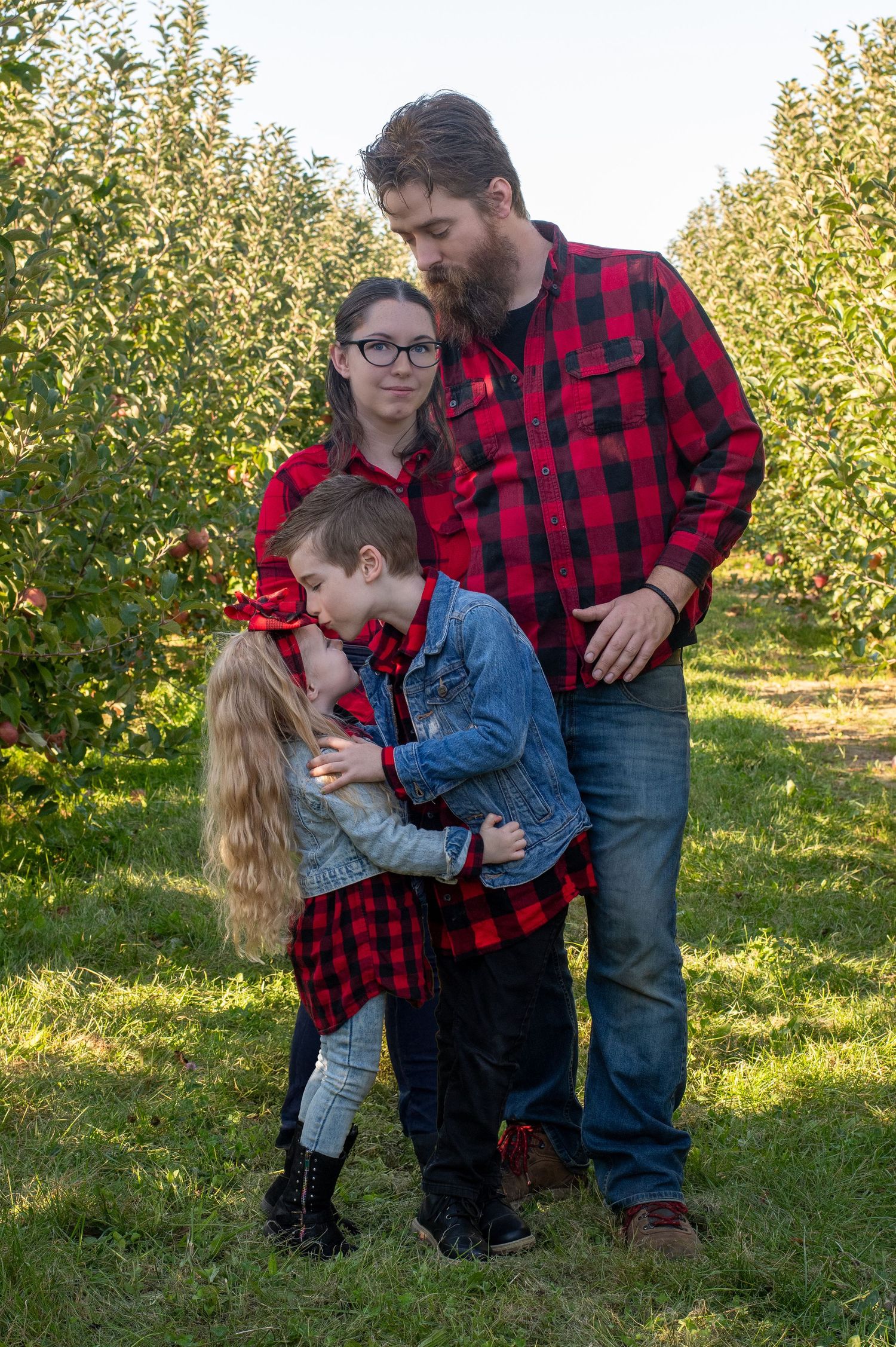 Family wearing matching buffalo plaid shirts poses together in apple orchard.