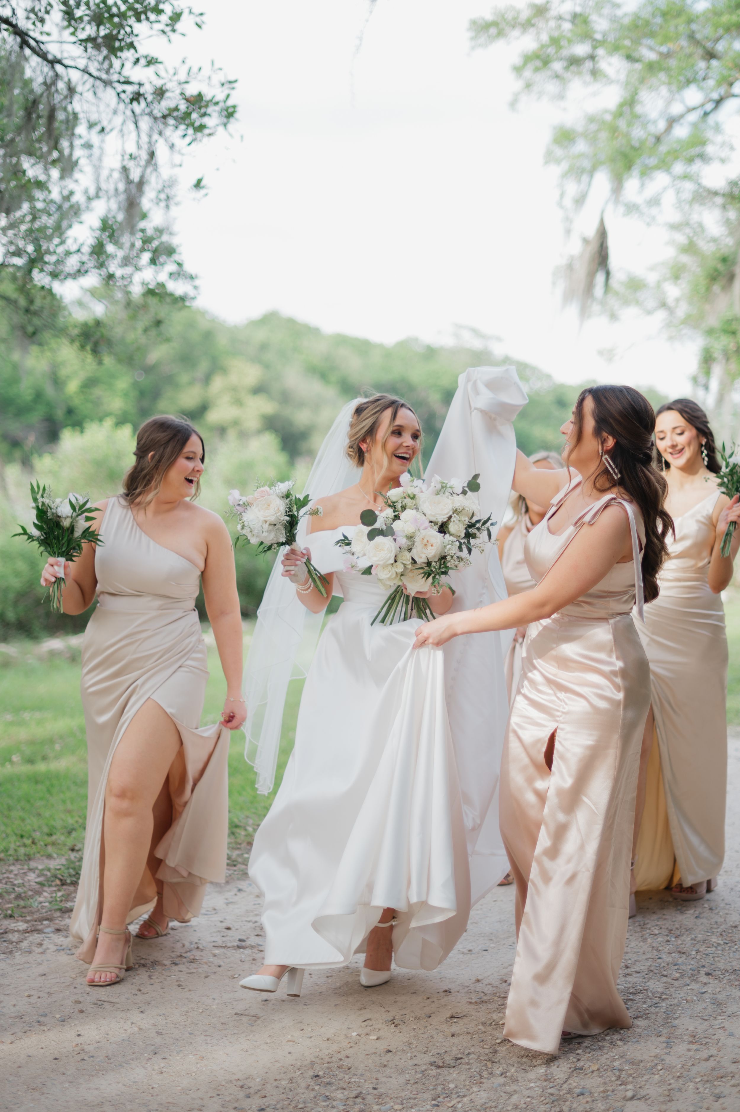 A wedding party walking outdoors on a dirt path with bridesmaids in light cream dresses and bouquets in nature setting.
