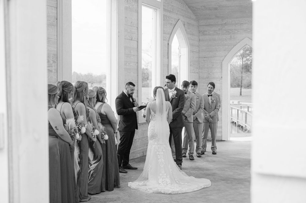 Black and white photograph of a wedding ceremony inside a bright church with bridal party members lined up on both sides.