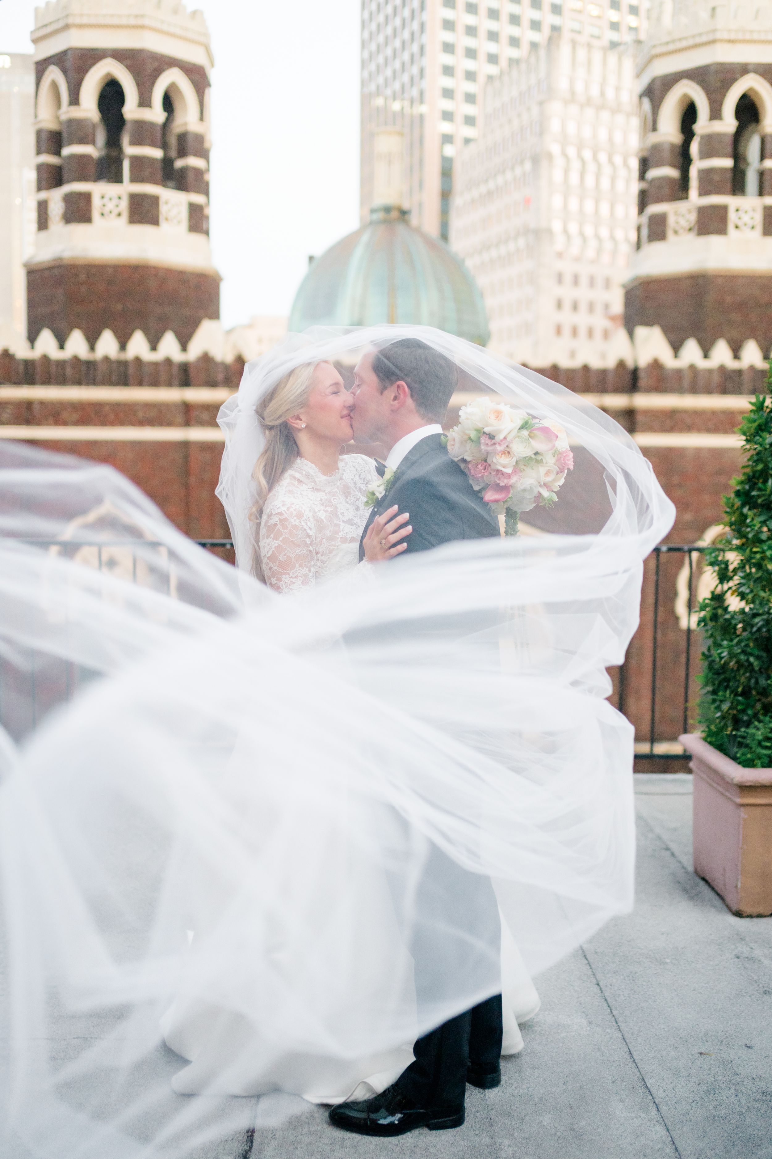Bride and groom kissing under flowing veil on rooftop with historic church towers in background.
