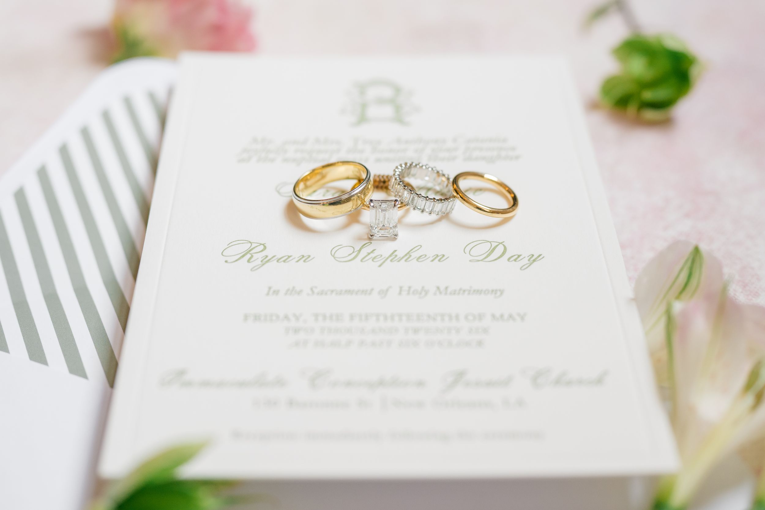 Gold wedding rings rest on an elegant white wedding invitation for Ryan Stephen Day, surrounded by soft floral accents.