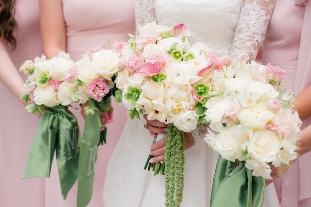 Bride and bridesmaids holding elegant white and pink floral bouquets with green ribbon at a wedding.