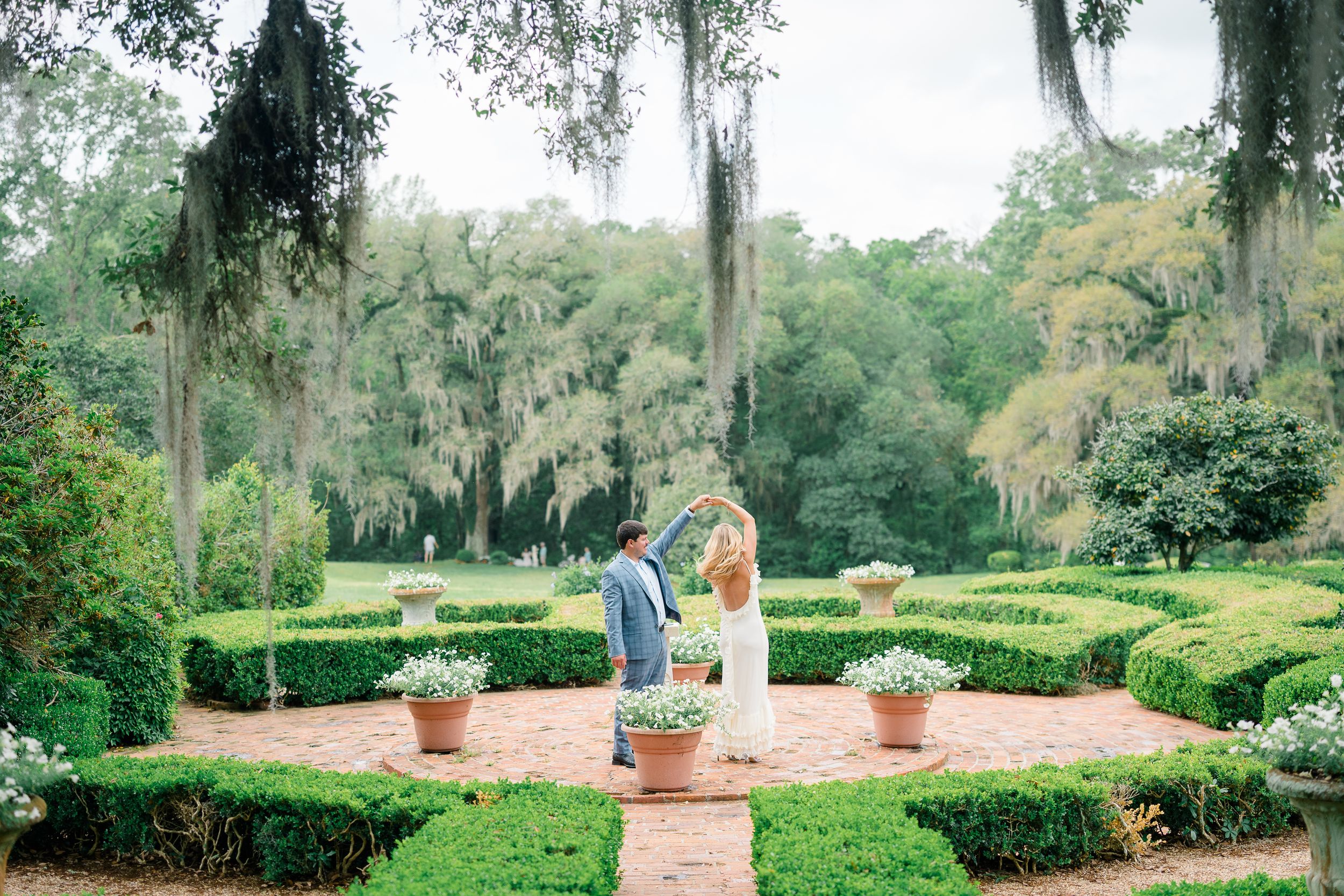Couple dancing in a lush formal garden with boxwood hedges, brick pathways, and Spanish moss-draped trees.