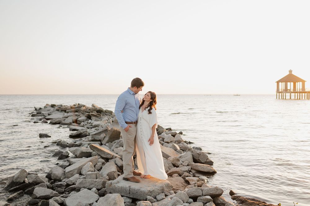 A romantic couple stands on a rocky jetty at sunset with a pier gazebo visible in the distance at the beach.