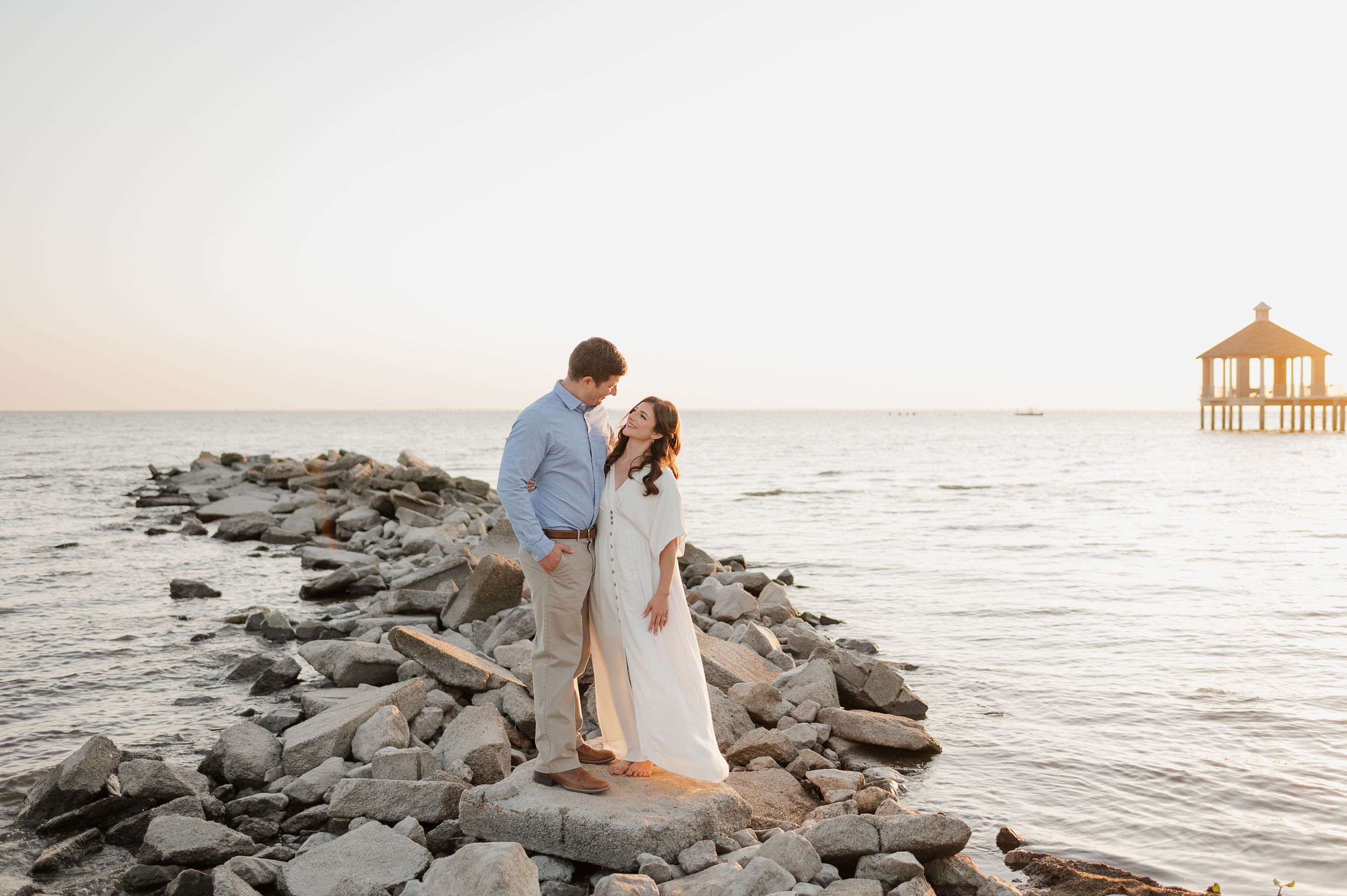 A romantic couple stands on a rocky jetty at sunset with a pier gazebo visible in the distance at the beach.