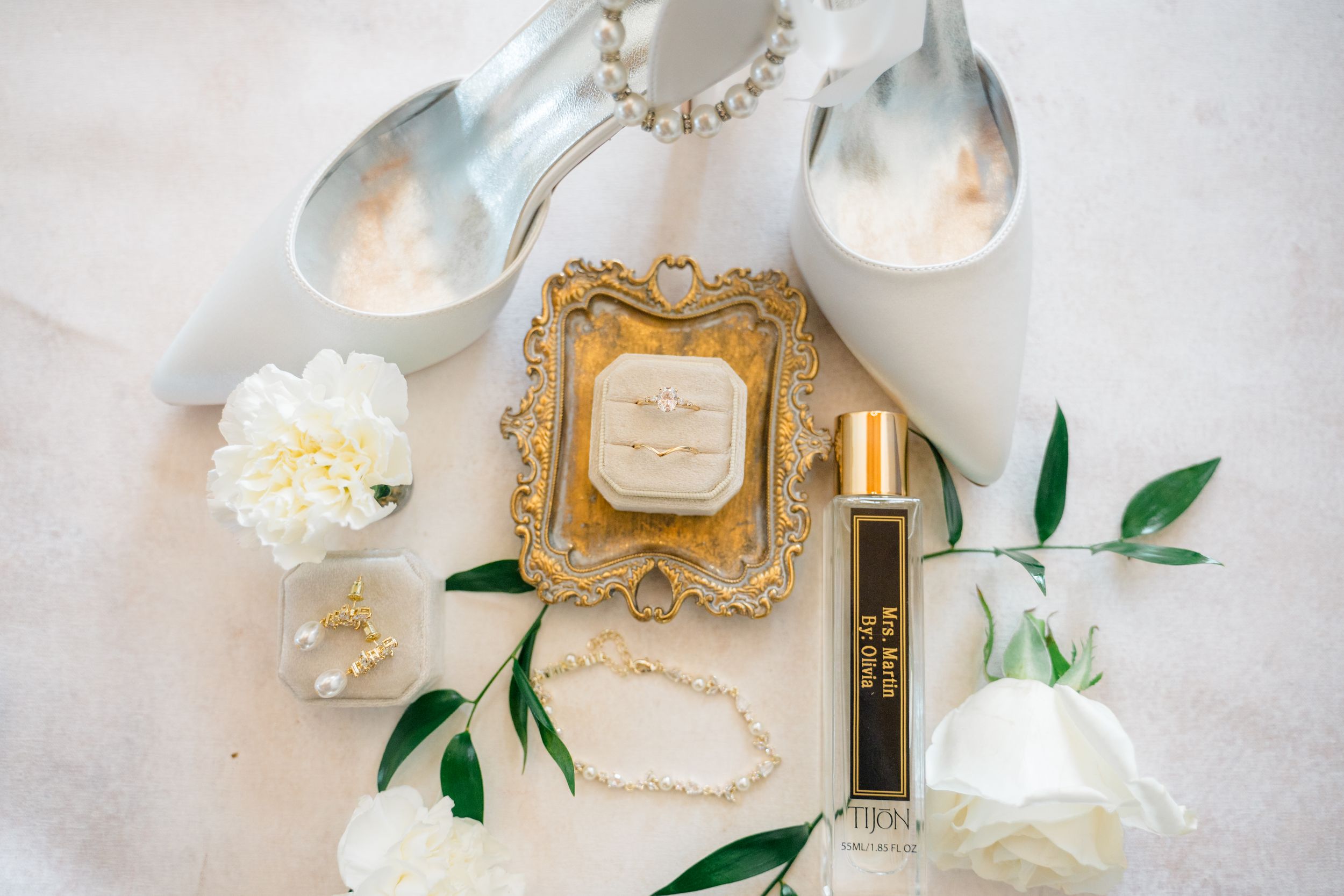 Elegant wedding flat lay with gold ring box, perfume, pearl bracelet, white roses, and bridal heels on soft white background.