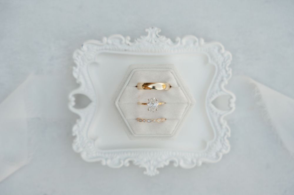 Two gold wedding rings displayed on a white hexagonal dish with ornate baroque frame against light fabric background.
