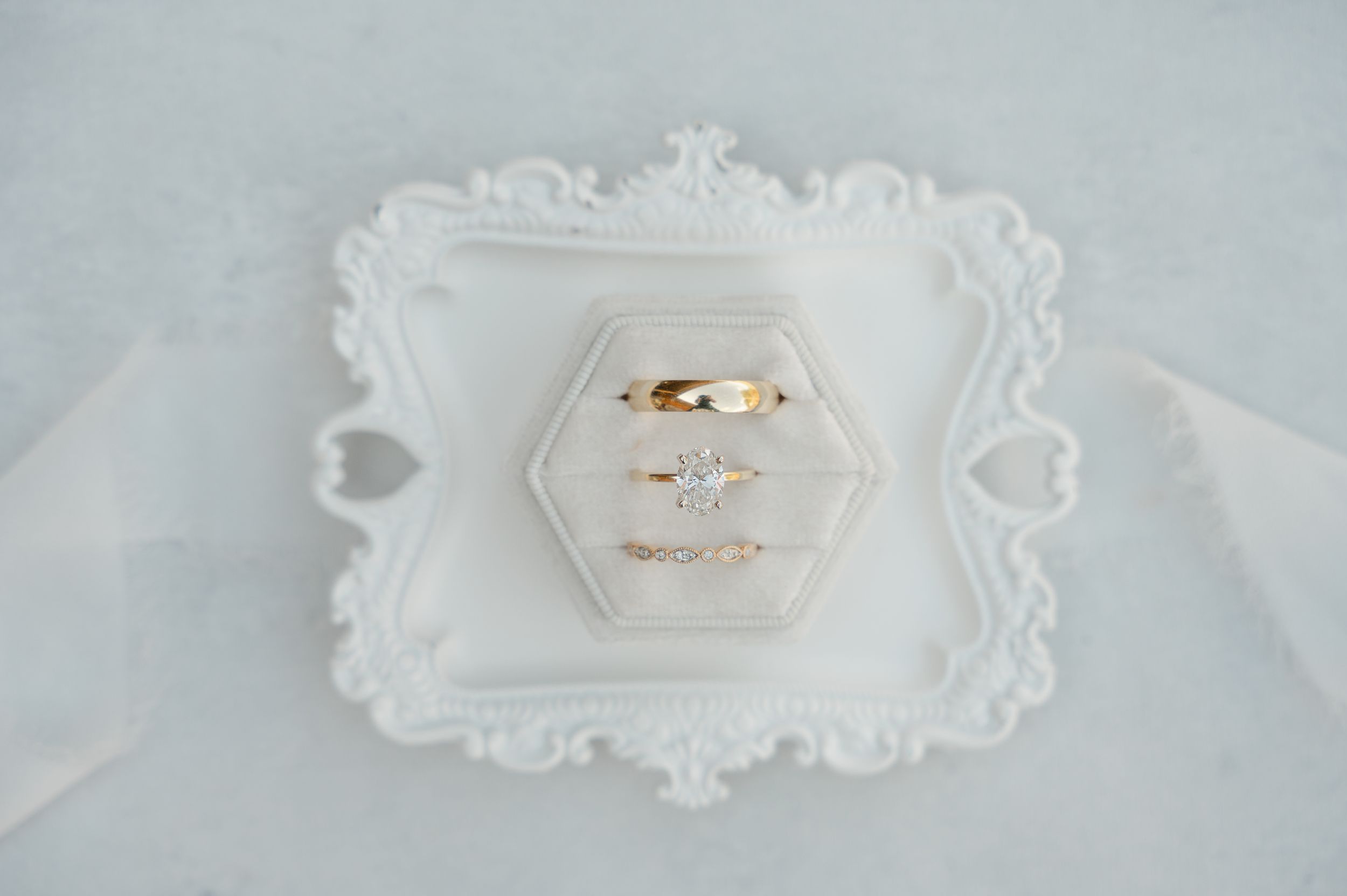 Two gold wedding rings displayed on a white hexagonal dish with ornate baroque frame against light fabric background.