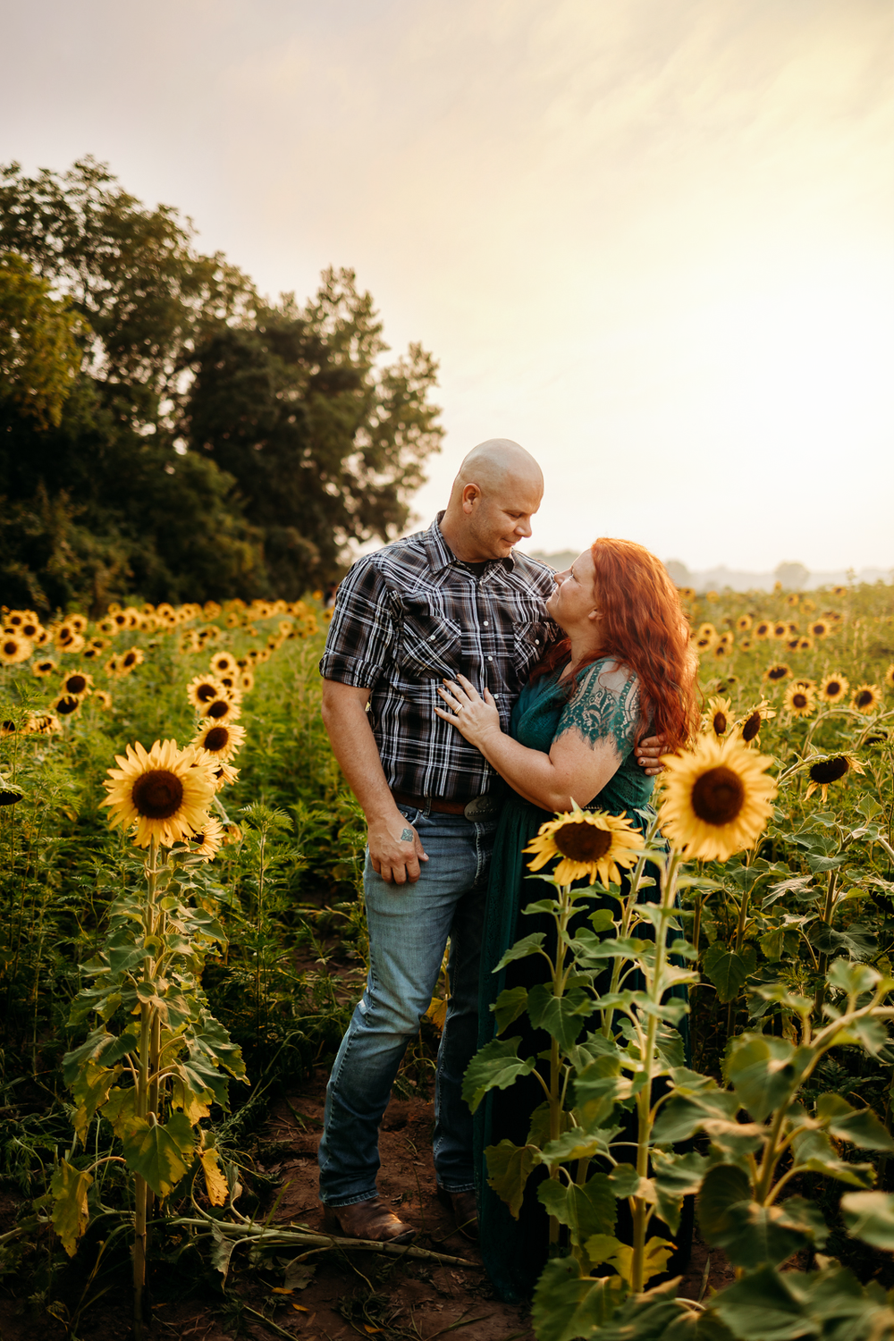 A couple lovingly gazing at each other in a field of sunflowers with a sunset in the background.