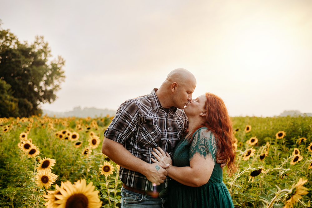 A couple kissing each other in a field of sunflowers with a sunset in the background.