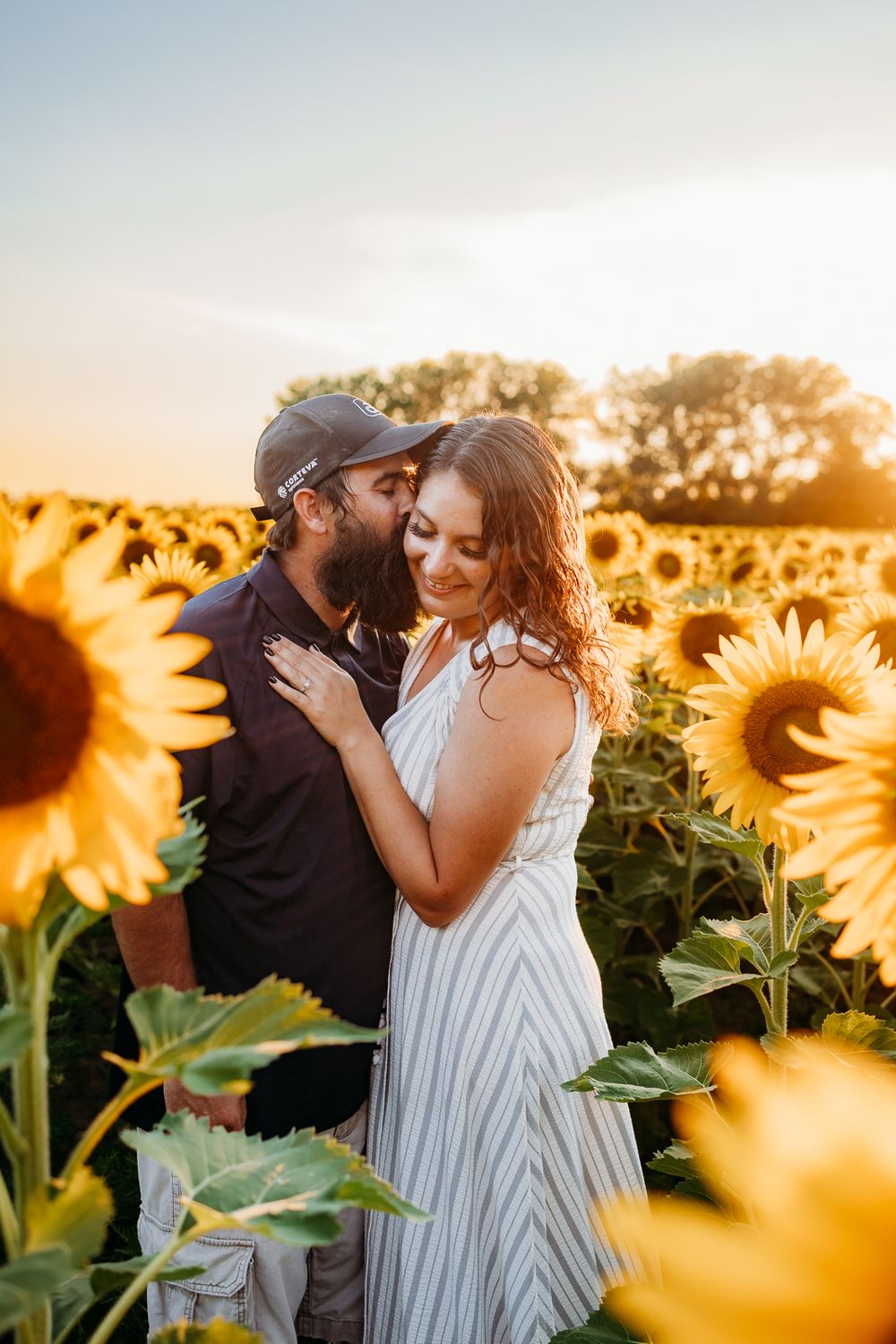 A husband and wife caressing each other in a field of sunflowers. The husband is kissing her on the cheek, while she smiles.