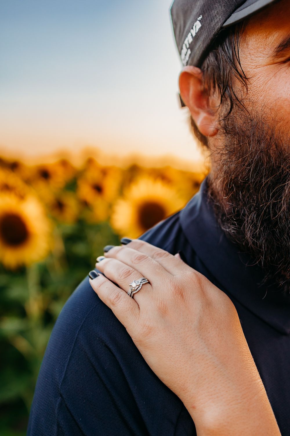 A wife's hand with her ring resting on her husband's chest in a field of sunflowers.