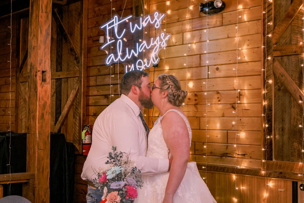 Wedding couple kisses under rustic wooden beams with glowing neon sign and string lights creating romantic ambiance.