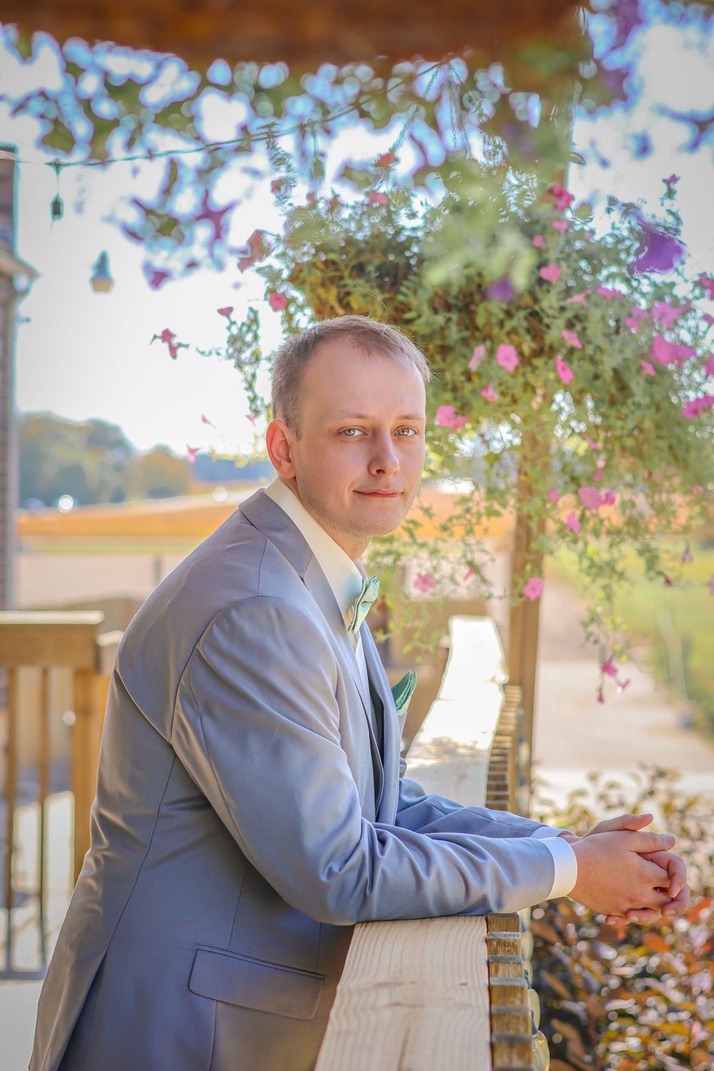 Person in light gray suit poses on porch with flowering plants in background.