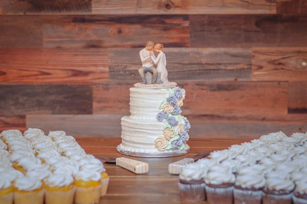 White wedding cake with blue floral decorations surrounded by cupcakes on rustic wooden background.
