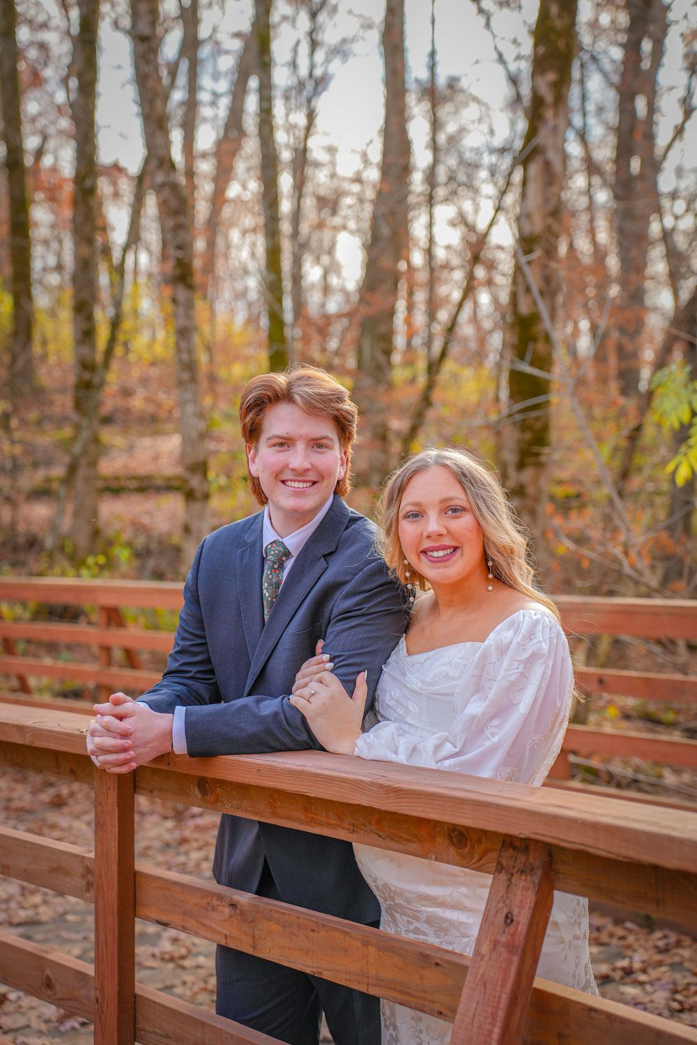 Wedding couple smiles while posing on wooden bridge in autumn forest.