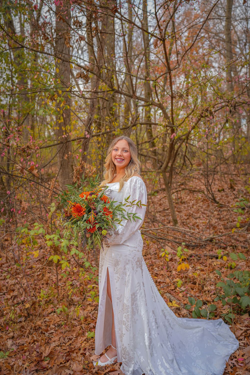 Bride in white lace gown holding colorful fall bouquet poses among autumn trees and fallen leaves.