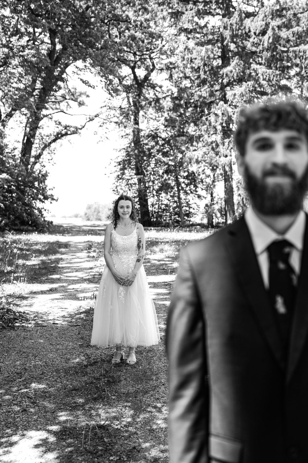 Black and white wedding photo of a bearded groom in foreground with bride standing on a tree-lined path behind him.