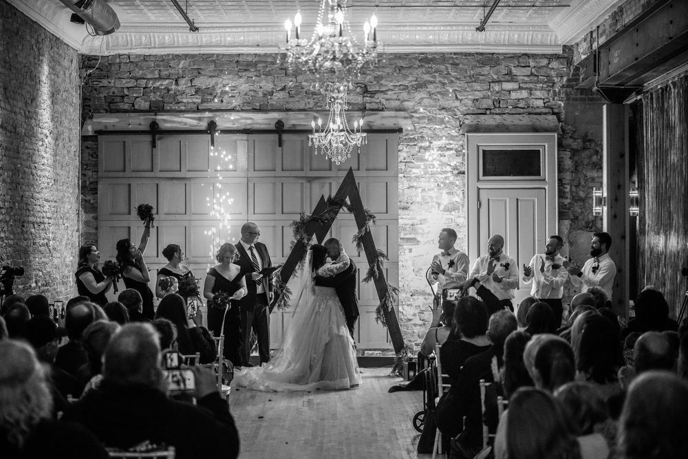 A black and white wedding ceremony takes place under a rustic wooden arch with chandelier in a brick-walled venue.