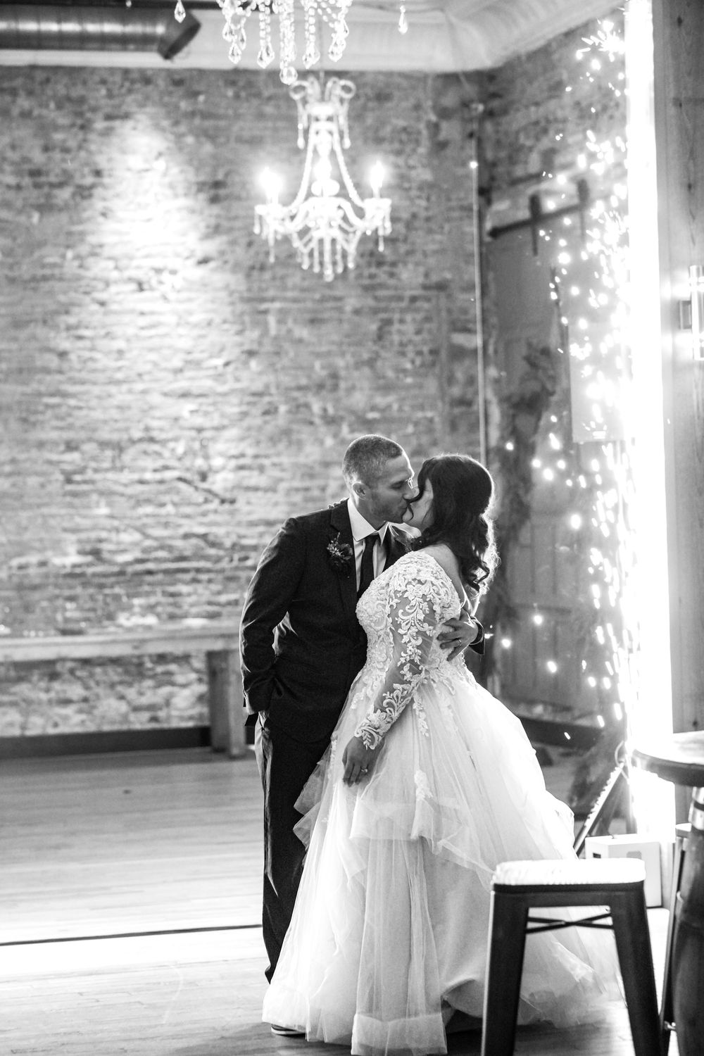 A romantic black and white wedding scene showing a couple dancing in an elegant brick-walled venue with sparkling chandeliers.
