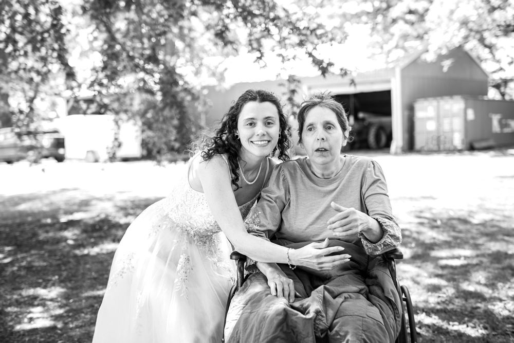 Bride in white gown embraces elderly woman in wheelchair outdoors in black and white wedding photo.