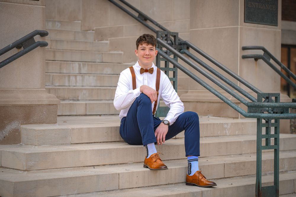 Young man in white shirt, suspenders, bow tie, and brown loafers sitting on stone steps for a senior portrait.