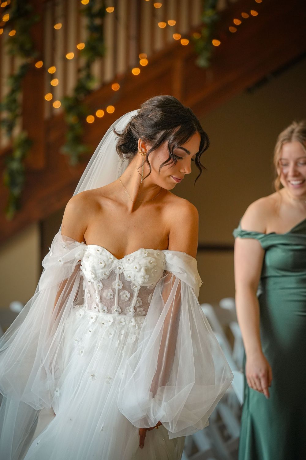 Bride in strapless lace tulle wedding gown with veil, standing beside smiling bridesmaid in green dress.