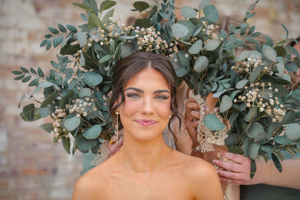 Bride with natural makeup poses with large eucalyptus and berry bouquet, wearing gold earrings against rustic brick wall.
