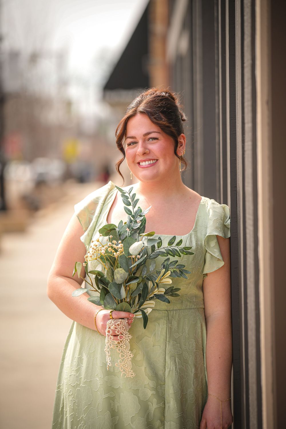 Smiling bridesmaid in sage green dress holding bouquet of eucalyptus and white flowers outdoors.
